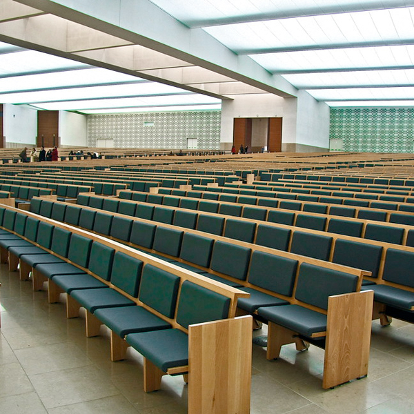 Un auditorium spacieux et moderne avec des rangées de bancs en bois du Banc FÁTIMA avec des coussins verts, de hauts plafonds et des lumières vives au plafond ; quelques personnes sont visibles près de l'entrée à l'arrière-plan.
