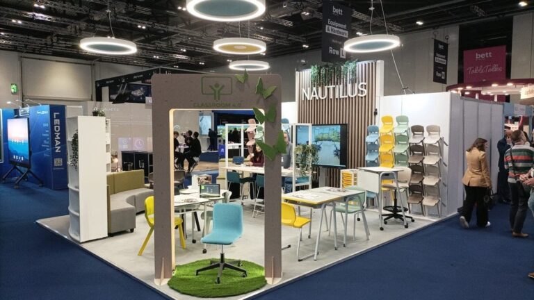 Un stand d'exposition moderne présente des meubles de classe colorés, des chaises et des bureaux bien rangés. Un éclairage décoratif est suspendu au-dessus, avec un panneau indiquant Nautilus à l'avant-garde de l'éducation - faits marquants du BETT Londres 2024 et des accents écologiques verts.