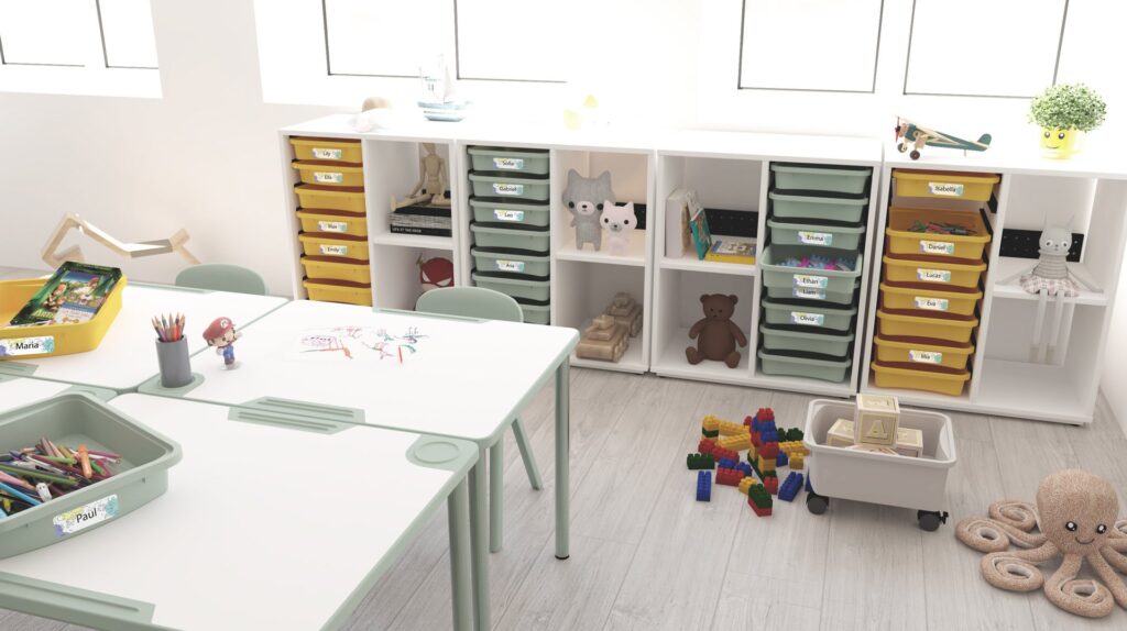 Une salle de classe lumineuse et organisée avec des étagères blanches contenant des tiroirs jaunes et verts étiquetés, des jouets, des livres et des animaux en peluche. Des tables contiennent des fournitures artistiques et des dessins, tandis que des blocs de construction et des ressources de l'école numérique sont éparpillés sur le sol en bois clair.