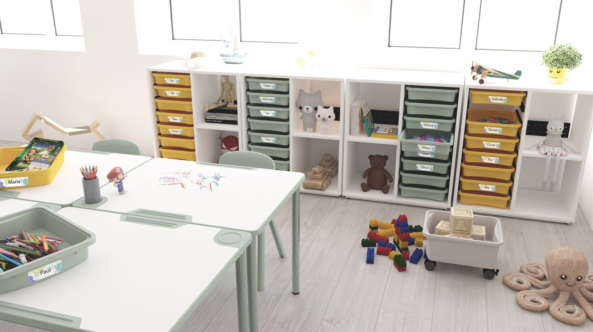 Une salle de classe lumineuse et organisée avec des étagères blanches contenant des tiroirs jaunes et verts étiquetés, des jouets, des livres et des animaux en peluche. Des tables contiennent des fournitures artistiques et des dessins, tandis que des blocs de construction et des ressources de l'école numérique sont éparpillés sur le sol en bois clair.