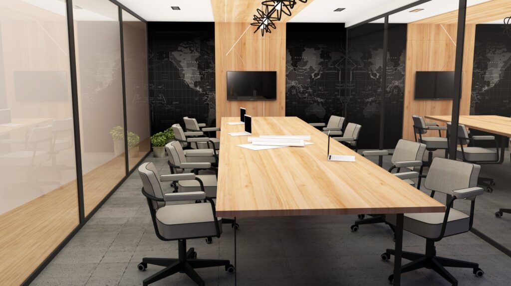 Salle de conférence moderne avec une grande table en bois, plusieurs chaises à roulettes, des téléviseurs au mur, des papiers et des stylos éparpillés sur la table, des cloisons en verre et une carte du monde sombre sur le mur du fond.
