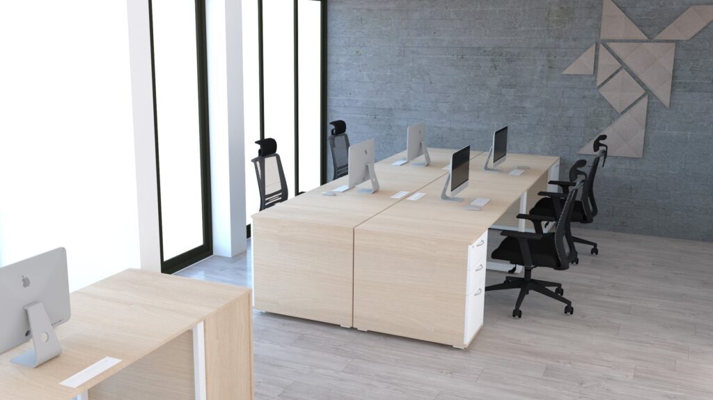 Un bureau moderne avec des bureaux en bois clair, des chaises ergonomiques noires et plusieurs ordinateurs de bureau. De grandes fenêtres laissent entrer la lumière naturelle et des œuvres d'art géométriques décorent un mur gris. L'espace semble propre et organisé.