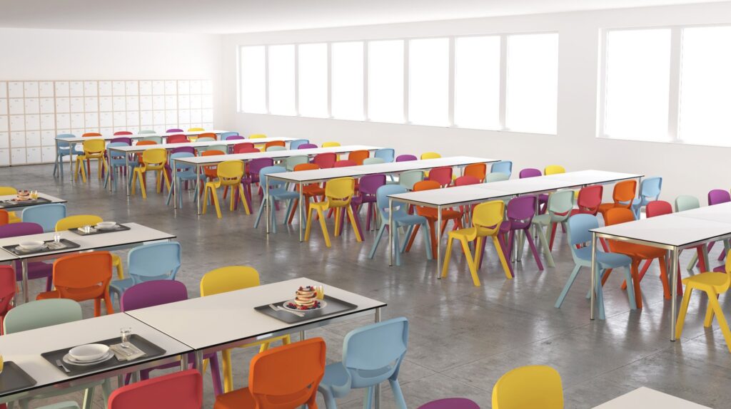 Une cafétéria lumineuse avec des tables blanches et des chaises colorées en orange, violet, rouge et jaune. L'espace est bien éclairé par de grandes fenêtres et des casiers bordent le mur du fond. Quelques plateaux contenant de la nourriture et de la vaisselle sont posés sur les tables.