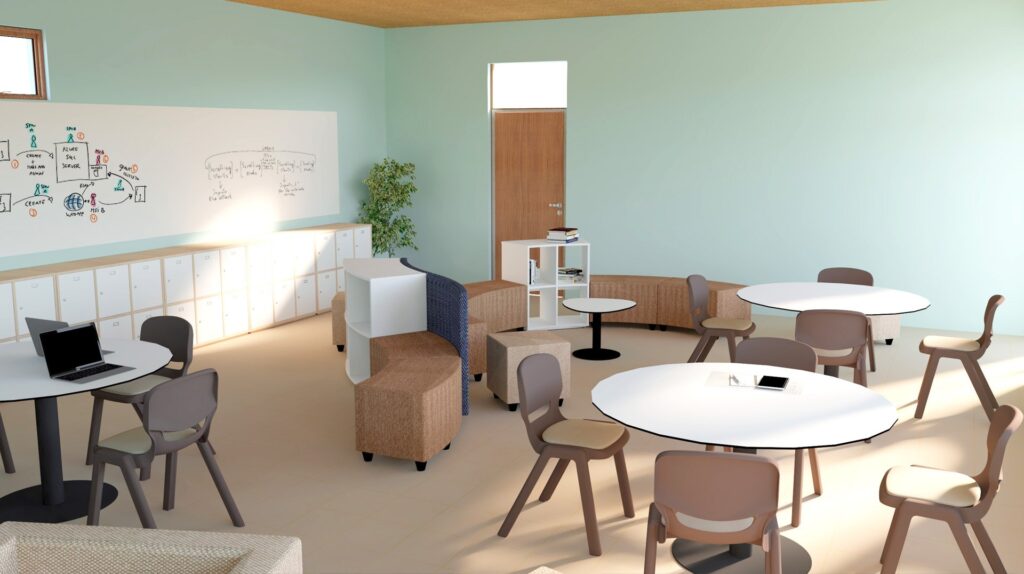 Une salle de classe moderne avec des tables rondes, des chaises, un tableau blanc avec des diagrammes, des casiers le long du mur, un ordinateur portable sur une table et une petite plante près d'une porte fermée ressemble à une salle des enseignants spacieuse avec des murs pastel et beaucoup de lumière naturelle.