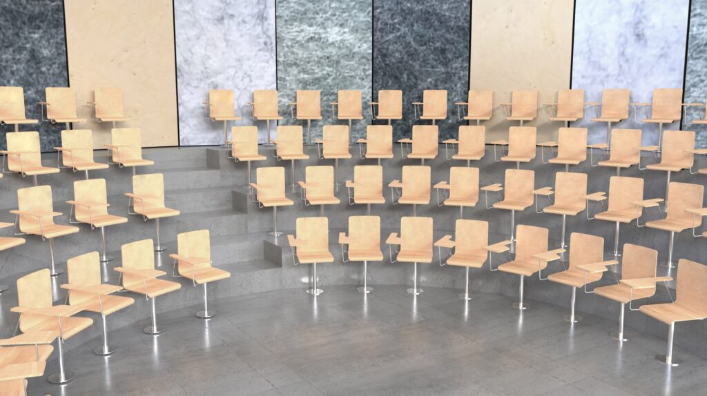 Un amphithéâtre moderne et vide avec des chaises en bois clair disposées en rangées courbes et étagées. Le sol est en béton gris et les murs sont constitués de grands panneaux de marbre beige et gris texturé.