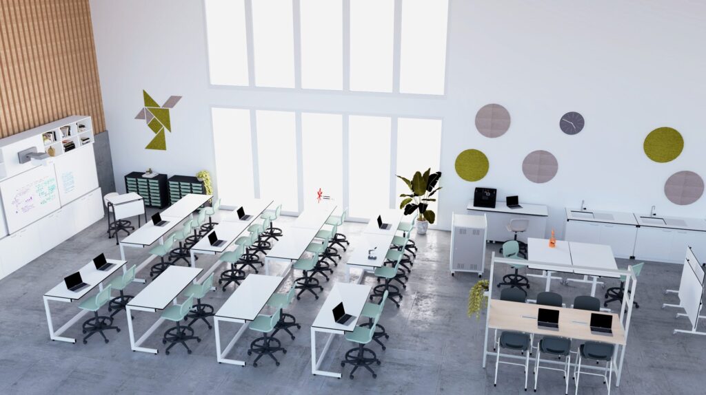 Salle de classe moderne avec des rangées de bureaux et de chaises blancs, chacun équipé d'un ordinateur portable. De grandes fenêtres inondent l'espace de lumière naturelle. Les murs sont couverts de plantes, d'œuvres d'art et de tableaux blancs, et les espaces de travail collaboratif ressemblent à des laboratoires d'apprentissage.