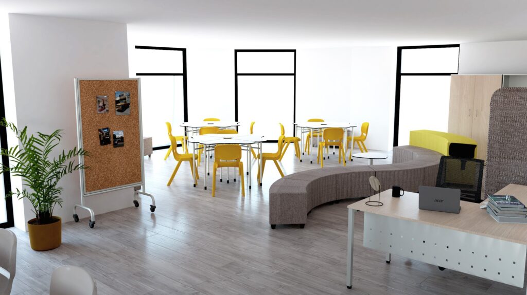 Salle de classe moderne avec de grandes fenêtres, des chaises jaunes autour de tables rondes, un tableau de liège sur roulettes, un canapé gris incurvé et un bureau blanc avec un ordinateur portable et des livres - comme une mini-bibliothèque - ainsi qu'une plante verte sur un sol en bois clair.