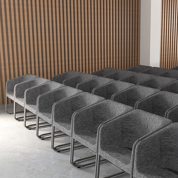 Des rangées de chaises modernes, grises et rembourrées, avec des pieds en métal noir, sont soigneusement disposées dans une pièce au sol en béton et aux murs en lattes de bois. Il semble s'agir d'un espace de conférence ou de réunion.