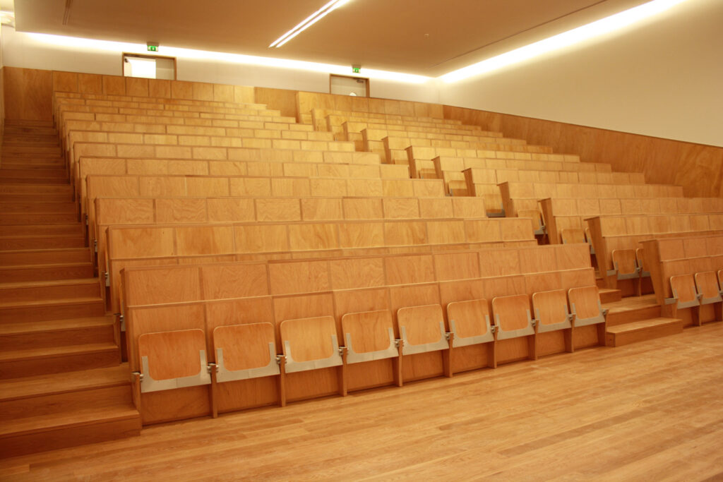 Un amphithéâtre vide avec des gradins en bois et un plancher en bois assorti, éclairé par des plafonniers et doté de sorties à l'arrière, pourrait inspirer mobilier de bureau ou même servir de toile pour mobilier innovant.