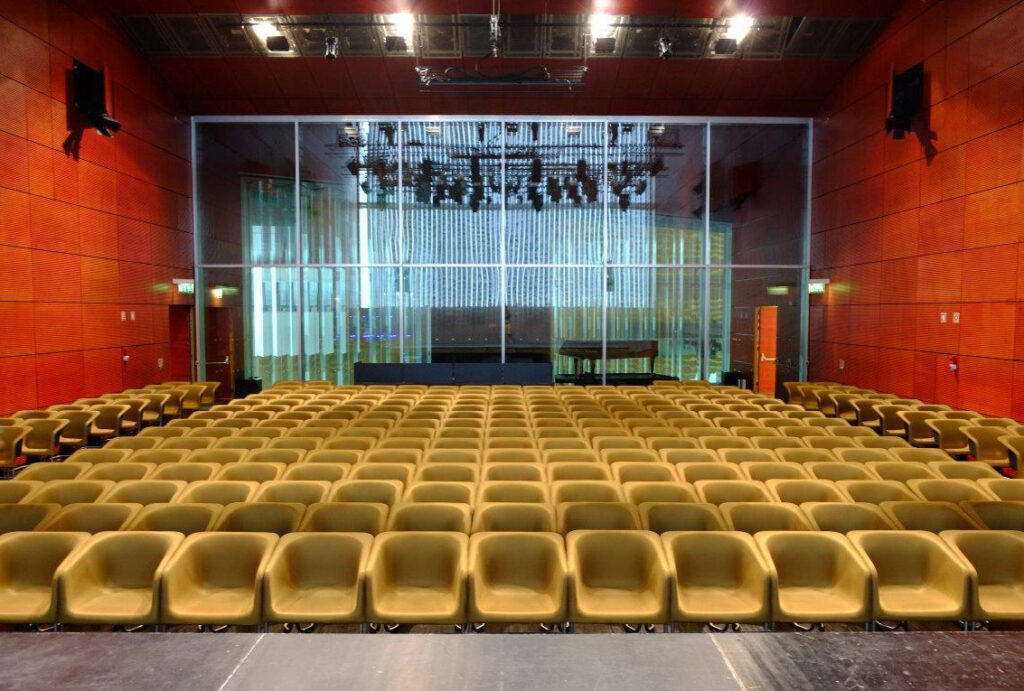 Un auditorium vide avec des rangées de chaises jaunes comme mobilier de bureau fait face à une scène, des murs lambrissés et une grande baie vitrée derrière la scène révélant l'équipement d'éclairage et un piano à queue.