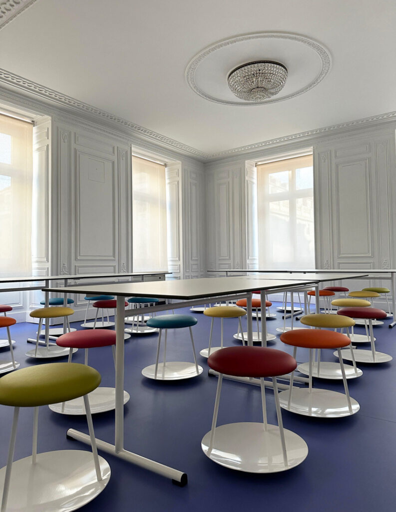 Une pièce lumineuse et élégante aux murs blancs ornés et aux grandes fenêtres présente des rangées de tables et des tabourets ronds colorés sur un sol bleu, mettant en valeur le mobilier de bureau innovant pour une atmosphère moderne et accueillante.