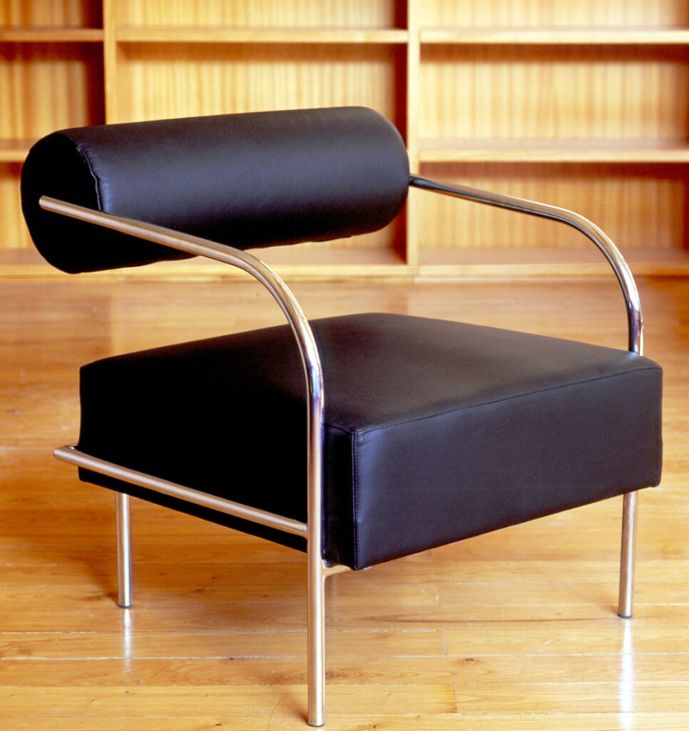 Une chaise moderne en cuir noir avec un dossier cylindrique et une structure métallique élégante est posée sur un sol en bois poli, mettant en évidence le mobilier de bureau innovant, avec des étagères en bois vides à l'arrière-plan.