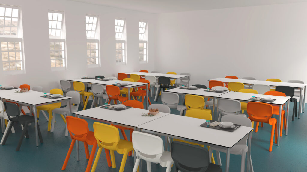 Une cafétéria lumineuse aux murs blancs et aux grandes fenêtres, meublée de tables rectangulaires et de chaises en plastique colorées en jaune, orange et blanc. Sur certaines tables sont disposés des plateaux, des assiettes et des ustensiles.