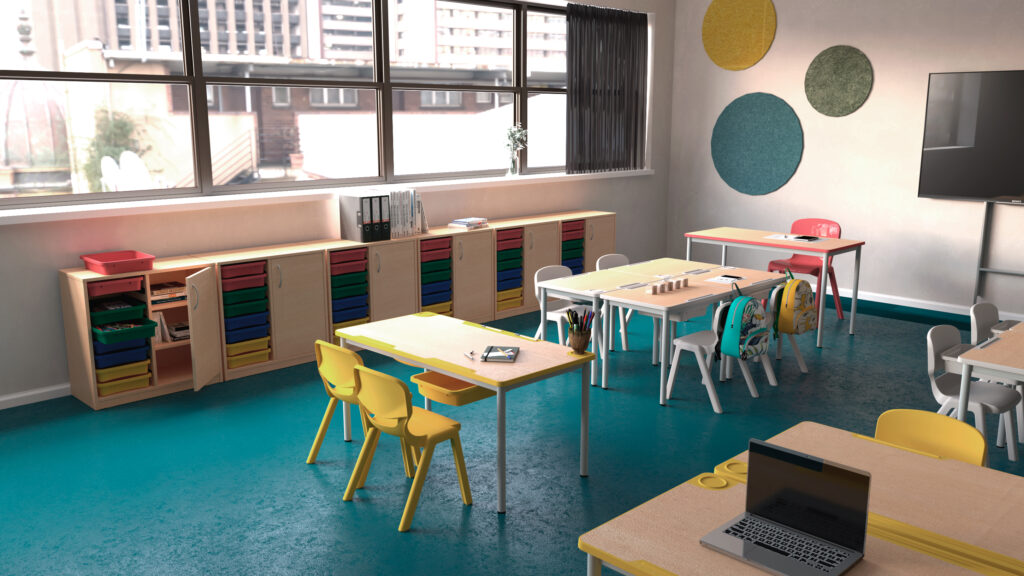 Une salle de classe lumineuse avec des chaises et des tables colorées, des armoires de rangement avec des bacs, un ordinateur portable sur un bureau et de grandes fenêtres laissant entrer la lumière naturelle. La décoration comprend des œuvres d'art murales circulaires et des classeurs sur les armoires.